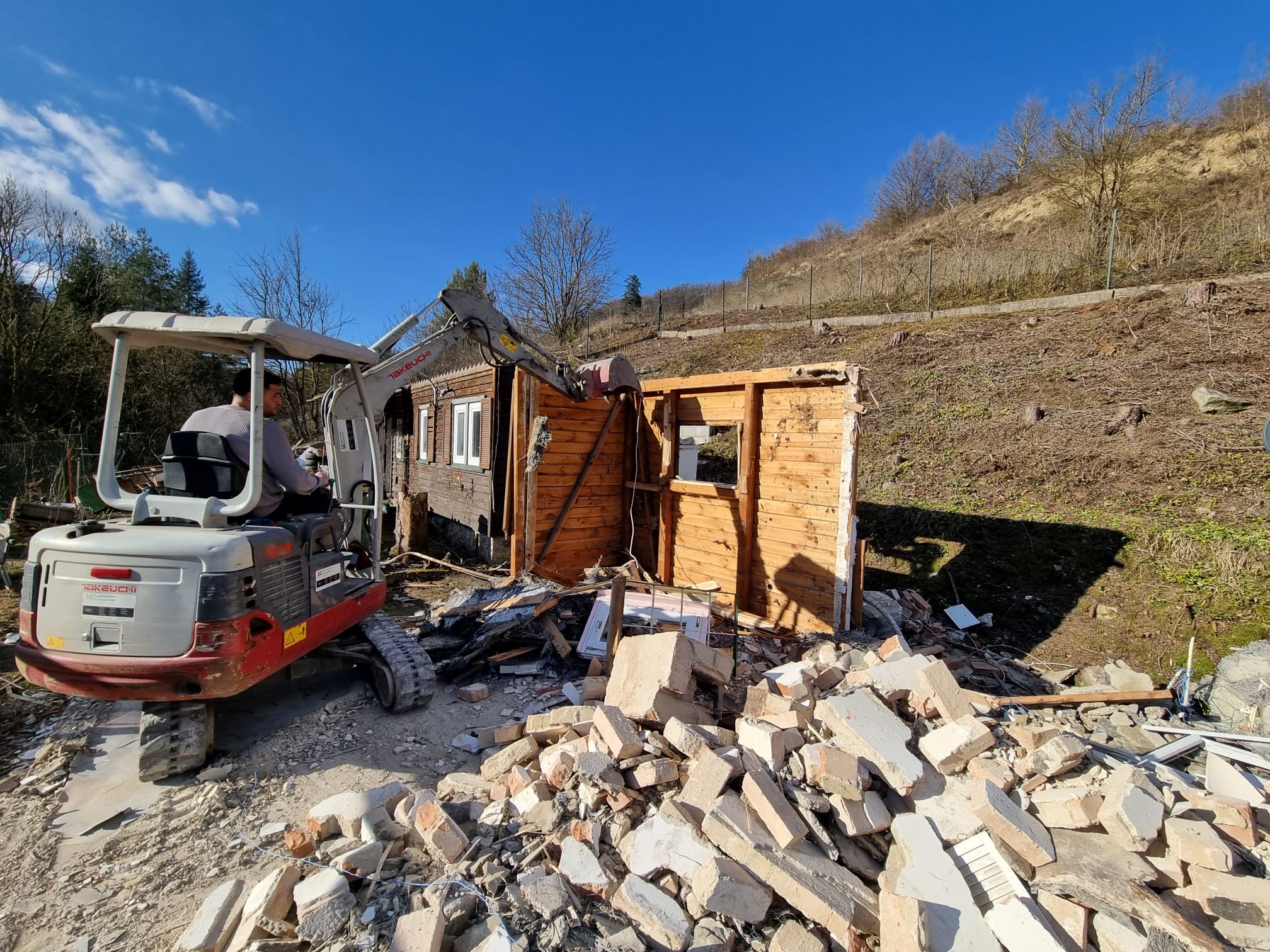 Fahrer im Takeuchi-Minibagger beim Abbruch eines Kleingartenhauses mit Ziegelschutt im Vordergrund